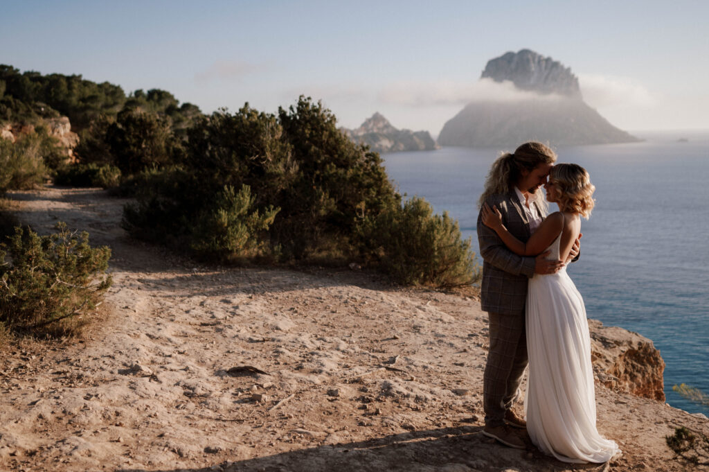 Couple standing on a cliff and holding each other during their Ibiza wedding shoot, with the sea and mountains in the background