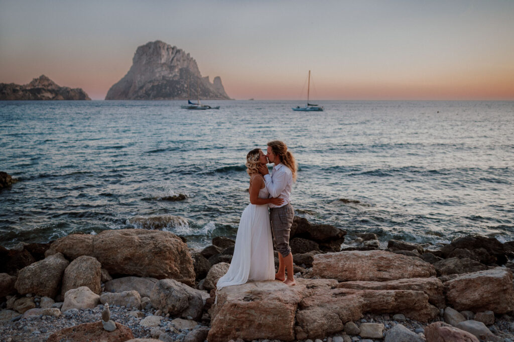 Wedding couple holding each other and kissing on a rocky beach in Ibiza during sunset